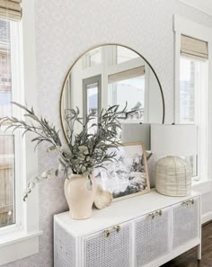 a white dresser topped with a mirror and vase filled with flowers next to a window