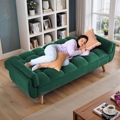 a woman laying on top of a green couch in a living room next to a window