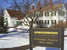 a sign in front of a large white house with snow on the ground and trees