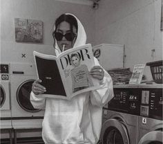 a woman in a hoodie reading a newspaper while standing next to washers and dryer machines