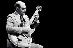 a black and white photo of a man holding a guitar in his right hand while wearing a suit