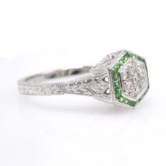 Made of 14K white gold, a cluster of 7 diamonds is surrounded by a hexagonal halo of scissor cut trapezoidal green (tsavorite) garnet. This stunning Art Deco style ring offers significant bang for the buck... and these diamonds absolutely dazzle. The sides of the ring have an engraved floral motif accented by dramatic geometric cut outs. Metal: 14K White Gold Main Stones: 7 Round Brilliant Diamonds (approx. 0.17ct total, G-H color, SI clarity) Ring Size: 6.5 (can be sized) Art Deco Green Emerald Ring With Rose Cut Diamonds, Art Deco Emerald Ring With Rose Cut Diamonds, Elegant Green Octagon Diamond Ring, Green Asscher Cut Diamond Ring, Octagon Emerald Ring With Halo Setting, Green Octagon Diamond Cut Ring, Green Art Deco Diamond Ring With Diamond Cut, Art Deco Diamond Emerald Ring Octagon Shape, Heirloom Green Diamond Ring With Diamond Cut