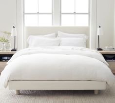 a bed with white linens and pillows in a bedroom next to two large windows