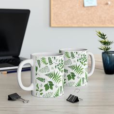 two coffee mugs sitting next to each other on a table with a laptop in the background