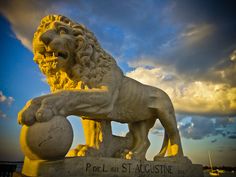 a statue of a lion holding a ball in front of a cloudy blue and yellow sky