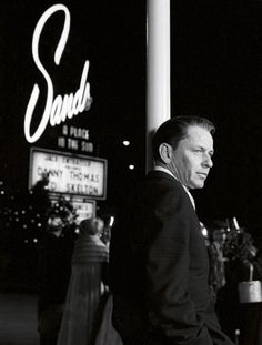 black and white photograph of man standing in front of sands casino sign at night time