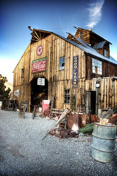 an old wooden building sitting on top of a gravel lot next to a metal barrel