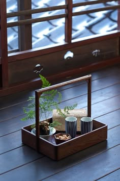 a wooden tray with two cups and a plant in it sitting on a wood floor