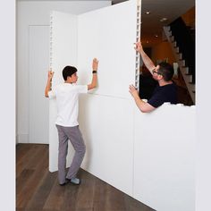 two men working on a wall in a room with wood floors and white walls, one holding his hands up to the wall while the other man is reaching for something