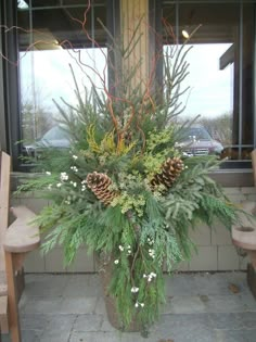 a vase filled with pine cones and greenery on top of a stone floor in front of a window