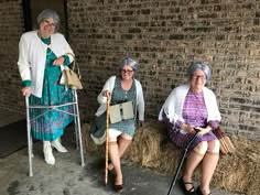 three older women sitting on hay bales in front of a brick building, one holding a crutch