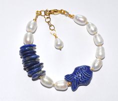 Indulge in the timeless beauty of our one-of-a-kind bracelet, meticulously crafted to captivate and enchant. Adorned with lustrous white freshwater pearls, mesmerizing lapis lazuli beads, and a charming porcelain fish, this piece is a celebration of elegance and sophistication. Lapis lazuli, a gemstone steeped in history and symbolism, takes center stage in this exquisite bracelet. Renowned for its stunning blue hue, flecked with golden pyrite, lapis lazuli has been cherished for millennia for its intense color and spiritual significance. Associated with wisdom and spirituality, it adds a touch of mystique and allure to any ensemble. Measuring approximately 18 cm (7") with an additional 3 cm extension chain adorned with a freshwater pearl, this bracelet offers both a perfect fit and a luxu Adjustable Blue Pearl Bracelet With Natural Stones, Adjustable Blue Pearl Bracelet With Gemstone, Blue Pearl Bracelet With Gemstone Beads For Gift, Blue Pearl Bracelet With Gemstone Beads As A Gift, Lapis Lazuli Bracelet With Natural Stones As Gift, Gift Bracelet With Lapis Lazuli Natural Stones, Lapis Lazuli Bracelets With Natural Stones As Gift, Lapis Lazuli Natural Stone Bracelet As A Gift, Lapis Lazuli Bracelet With Natural Stones