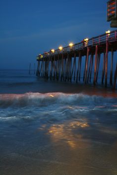 a long pier at night with lights on the water and waves coming in to shore