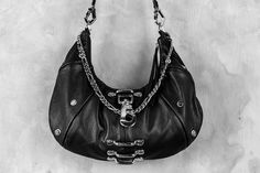 An ode to the timeless partnership of leather and chains, this leveled up black hobo bag is made from soft, premium genuine black leather and features a top shelf arrangement of silver hardware throughout.A centered, weighty metal industrial clip takes center stage, accompanied by an array of high contrast chromed out nailed rivets, centered feed through bars, and a draped top chain that doubles as a shoulder strap.Complete with a removable/adjustable leather and chain strap that can easily convert this purse from a shoulder bag to a comfortable crossbody bag, this design is an ode to the tough and timeless partnership of leather and chains.Bag is approx 13.5" x 7"Removable strap can extend up to 40" Formal Leather Hobo Bag With Gunmetal Hardware, Edgy Black Shoulder Bag With Palladium Hardware, Leather Tote Bag With Chain Strap, Edgy Leather Shoulder Bag With Metal Hardware, Leather Hobo Bag With Chain Strap For Daily Use, Black Edgy Bags With Palladium Hardware, Edgy Black Bags With Palladium Hardware, Leather Hobo Bag With Chain Strap, Leather Tote Shoulder Bag With Chain Strap