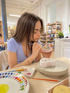 a woman sitting at a table painting on a plate with watercolors in front of her