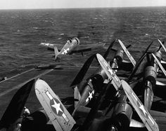 several planes are on the deck of an aircraft carrier in the ocean, while another plane flies overhead