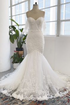 a white wedding dress on display in front of a window with potted plant behind it