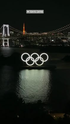 the olympic rings are lit up at night in front of tokyo's bay bridge