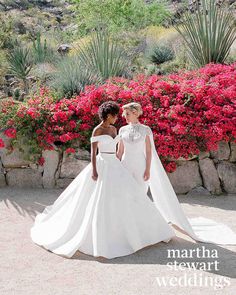 two women in wedding gowns standing next to each other near some bushes and flowers