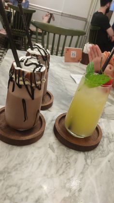 two drinks are sitting on wooden coasters at a table with people in the background