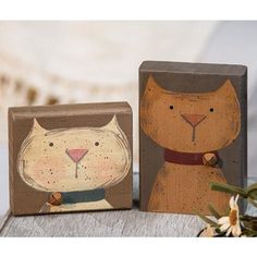 two wooden blocks with cats painted on them sitting next to each other and flowers in the background