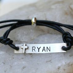 a black leather bracelet with a silver cross and the word ryan on it is sitting on a rock