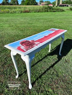 a white table with a red and blue painting on it in the middle of a grassy field