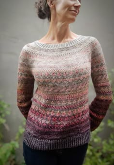 a woman wearing a multicolored sweater stands in front of a plant and looks off to the side