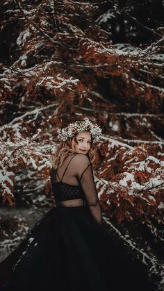 a woman wearing a black dress standing in front of snow covered trees with her hands on her hips