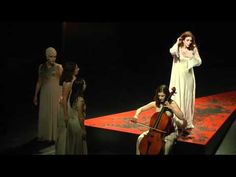 a woman in white dress playing violin on stage with other women standing around her and wearing long dresses