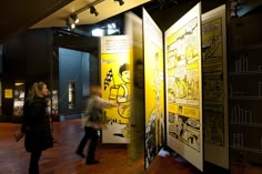 two people are walking by some posters in a room with wood flooring and black walls