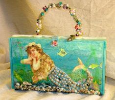 a handbag that has been painted with mermaids and pearls on the front, sitting on a white surface