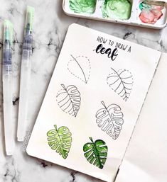 an open notebook next to some watercolors and pens on a marble surface with the words how to draw a leaf