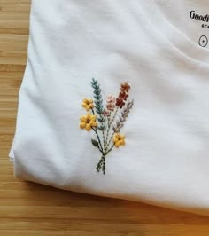 a white t - shirt with flowers embroidered on the front and back, sitting on a wooden surface