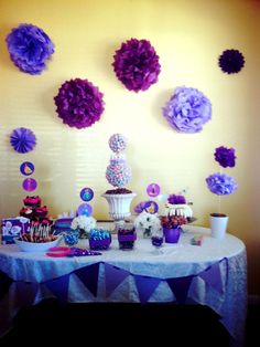 a table topped with lots of purple and white desserts next to a wall covered in paper flowers