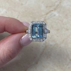 This stunning ring showcases a captivating 3.27 carats of emerald-cut aquamarine at its center. elegantly bezel-set and enhanced by a halo of old European cut diamonds; flanking the center stone are baguette-cut diamonds, adding to the overall allure. Each shoulder of the ring is adorned with a row of old Euro[pean cut diamonds. The total diamond weight of the ring is approximately 0.62 carats accentuating its brilliance.  The ring is handcrafted in platinum and features delicate milgrain detail Baguette Cut Topaz Ring With Diamond Halo Setting, Baguette Cut Topaz Ring With Diamond Halo, Gia Certified Emerald Cut Topaz Ring In Platinum, Gia Certified Emerald Cut Platinum Topaz Ring, Fine Jewelry Emerald Cut Topaz Ring Gia Certified, Heirloom Diamond Topaz Ring With Emerald Cut, Dazzling Emerald Cut Diamond Topaz Ring, Heirloom Diamond Topaz Ring, Emerald Cut, Emerald-cut Topaz Ring With Halo Setting