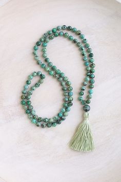 This mala necklace is made with 6-7mm African turquoise beads and a 10mm African turquoise guru bead. The beads are a green turquoise color with a brownish black matrix. Each strand of beads is unique. If you would like a photo of your mala before it is shipped out, please feel free to message me.  This mala is hand-knotted and made in the traditional way with 108 beads. It is 32" in length and the sage green tassel is available as either 2" or 3" long. The tassel is handmade using silk thread.  African turquoise encourages positive change, optimism and balance. It opens the mind to new ideas and helps us confidently walk towards our dreams. African turquoise is part of the jasper family. This mala will come in a stamped cotton bag and box with a card explaining the gemstone meaning. What Turquoise Healing Crystal Necklace With Round Beads, Turquoise Gemstone Beads Crystal Necklace, Adjustable Beaded Turquoise Necklace For Meditation, Adjustable Spiritual Turquoise Necklace For Meditation, Spiritual Turquoise Hand Knotted Jewelry, Spiritual Hand-knotted Turquoise Jewelry, Spiritual Turquoise Necklace With Round Beads For Meditation, Green Gemstone Beads Mala For Healing, Turquoise Spiritual Necklaces With 8mm Beads