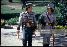 two men dressed in uniforms walking down a dirt road with trees and bushes behind them