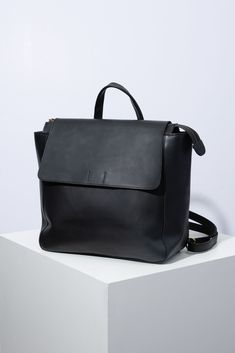 The Debut Backpack is a square leather bag with a wide base that allows it to sit upright. It is composed entirely of glossy black leather. There is a zipper closure across the entire bag. A black leather flap folds over the front of the bag, securing it further. The bag has a short black leather handle at the top, as well as a long, adjustable leather strap that allows the bag to be styled as a shoulder bag or backpack. Noonday Collection, Coffee Gifts Card, Commuter Bag, Black Leather Backpack, Coffee Gifts, Travel Pouch, Wallet Bag, Cute Bag, Summer Essentials