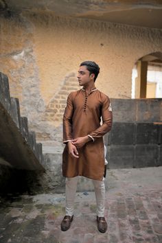 A brown silk kurta and fitted plain cotton silk pajama with gold embroidery on collar and sleeves and a matching motif on the arms, with round buttons to compliment the traditional look. Model wears a size Small. Ceremonial Cotton Silk Kurta With Long Sleeves, Ceremonial Churidar In Cotton Silk For Eid, Ceremonial Long Sleeve Cotton Silk Kurta, Ceremonial Long-sleeve Cotton Silk Kurta, Ceremonial Cotton Silk Churidar For Eid, Traditional Long Sleeve Cotton Silk Churidar, Long Sleeve Cotton Silk Kurta For Traditional Ceremonies, Ceremonial Long Sleeve Chanderi Kurta, Gold Cotton Silk Long Sleeve Kurta