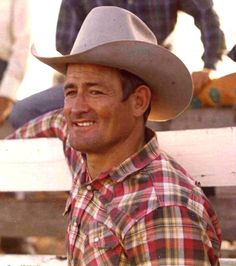 a man wearing a plaid shirt and cowboy hat smiles while sitting on a wooden bench