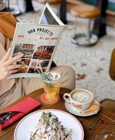 a woman sitting at a table reading a book and eating food with a cup of coffee in front of her