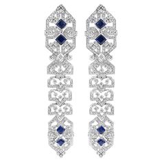 Stunning 18 karat white gold earrings adorned with diamonds and blue sapphires. The intricate design showcases a symmetrical pattern with princess-cut blue sapphires set in the centre, surrounded by sparkling diamonds. The geometric detailing adds a unique and luxurious touch, making these earrings a perfect statement piece for any elegant occasion. Diamond Total Weight: 6.4 carats All Mozafarian jewellery comes with a valuation certificate. Luxury White Gold Diamond Earrings With Intricate Design, Evening Sapphire Earrings With Diamond Accents, Evening Earrings With Sapphire And Diamond Accents, Sapphire Earrings With Diamond Accents For Evening, Luxury Platinum Blue Earrings, Luxury Blue Platinum Earrings, Elegant Blue Platinum Earrings, Luxury White Gold Sapphire Diamond Earrings, Formal Sapphire Earrings With Diamond Cut