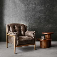 a brown chair sitting next to a wooden table on top of a cement floor with a gray wall behind it