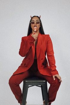 a woman in a red suit sitting on a stool with her tongue out and wearing glasses