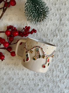 a coffee cup with christmas decorations on it next to a small pine tree and red berries