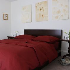 a bed with red sheets and pillows in a bedroom next to two pictures on the wall