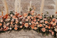 an arrangement of flowers and plants on the sand with feathers blowing in the wind behind them