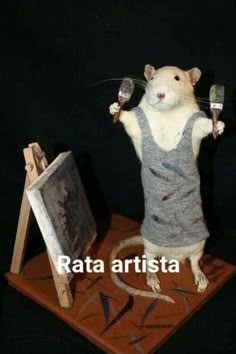 a rat holding two paintbrushes while standing on top of a wooden easel