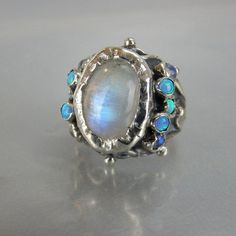 "A unique antique style hand crafted ring for brides to be! A rainbow moonstone ring that will make you feel like a real queen on your special day! This moonstone ring features a sterling silver band and handcrafted setting of fascinating rainbow moonstone gemstone, and small moonstones and lab opals stones on both sides. Measurements: Center stone - 10mm / 14mm Size: Moonstone queen ring is adjustable to any size, it has a small opening so you can adjust it to fit your finger. Materials: Stone: Mystical Cabochon Ring For Anniversary, Celestial Multi-stone Moonstone Ring, Celestial Multi-stone Moonstone Oval Ring, Celestial Multi-stone Oval Moonstone Ring, Mystical Anniversary Ring With Cabochon, Unique White Multi-stone Moonstone Ring, Moonstone Multi-stone Opal Ring As Gift, Multi-stone Moonstone Opal Ring For Gifts, Heirloom Style Moonstone Open Ring